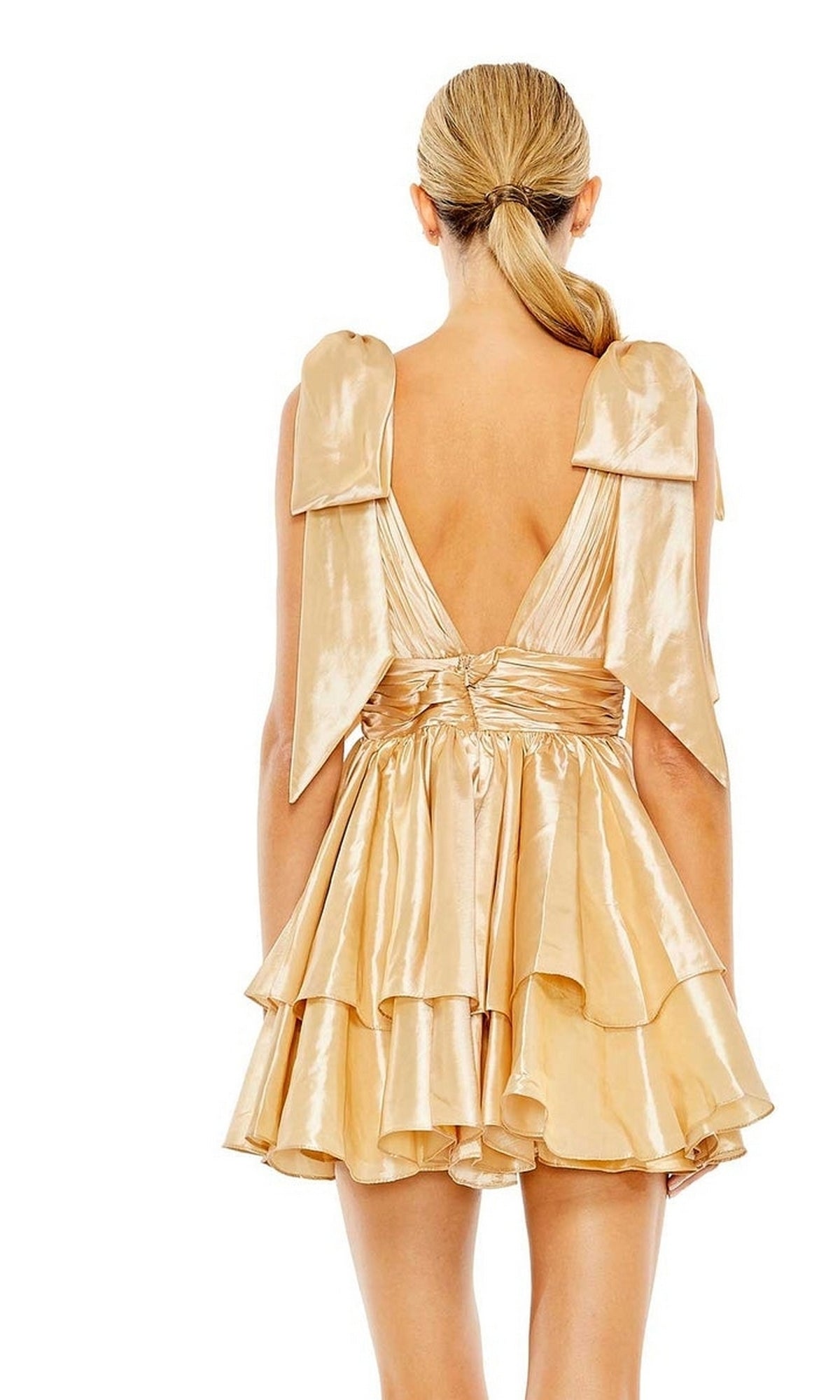 A blonde woman with a low ponytail models the Mac Duggal 11657 Bow Short Ruffled Hoco Dress, featuring shiny gold fabric, large shoulder bows, and a deep V-back, standing against a white background.